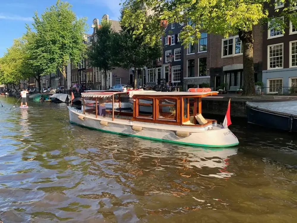 Small wooden-cabin canal boat with white hull on an Amsterdam canal beside tree-lined brick houses