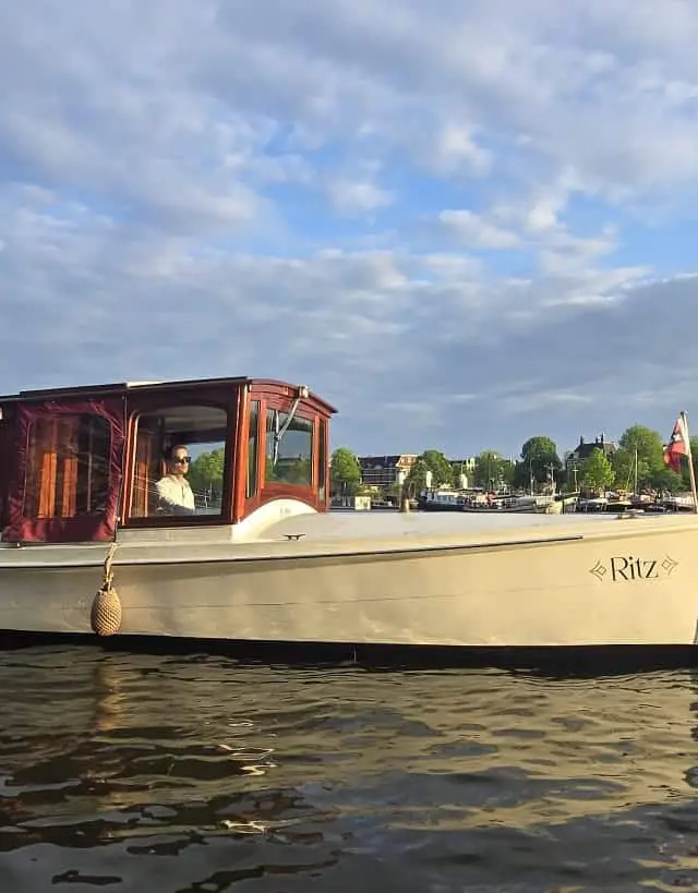 Ritz, a cream-hulled saloon boat with a red wooden cabin and skipper at the helm on Amsterdam water